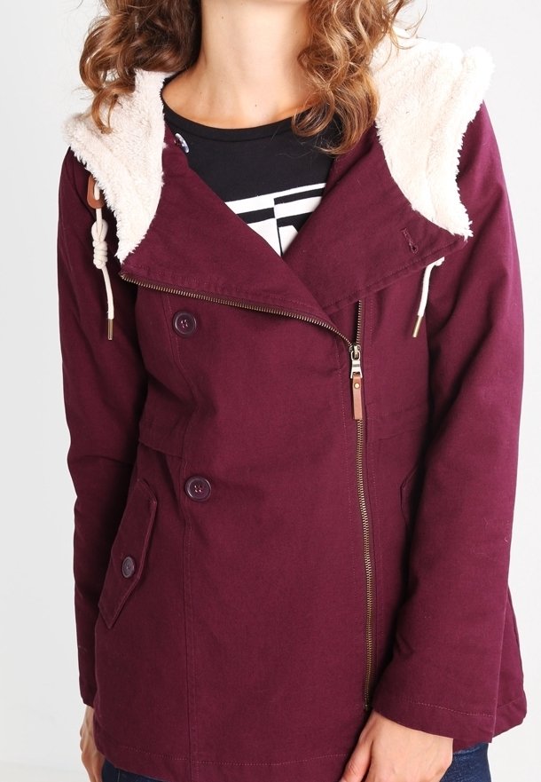 Femme portant une veste bordeaux avec capuche doublée en peau lainée, fermeture éclair diagonale, poche à rabat boutonnée et détails à boutons sur un haut noir.