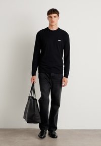 Schwarzes Langarmshirt mit kleinem weißen Logo, kombiniert mit dunklen Jeans und schwarzen Schuhen. Die Person hält eine schwarze Tragetasche.