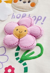 Fleur en tissu rose doux avec un centre jaune souriant et une tige verte cousue sur un fond blanc avec un texte violet "hop hop!"