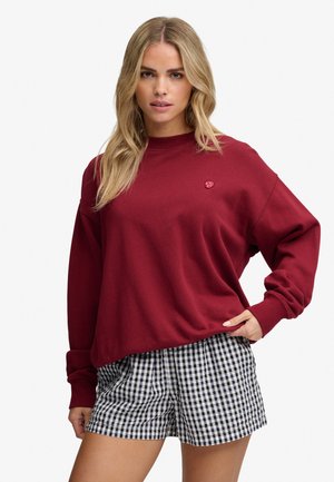 Blond kvinde iført en rød sweatshirt med lange ærmer og sort-hvide ternede shorts, stående med den ene hånd på hoften.