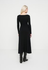 Black long-sleeve ribbed dress with a boat neckline, textured fabric, and a flowy silhouette, paired with knee-high black heeled boots.