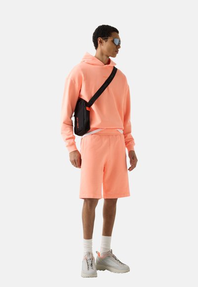 Mand iført koralrød hættetrøje og shorts, hvide sokker, beige sneakers, sort skuldertaske og reflekterende solbriller, stående mod hvid baggrund.