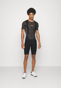 Bodysuit de sport noir à manches courtes avec sections en maille, design ajusté, accents gris et logo. Porté avec un short noir serré et des chaussures blanches.