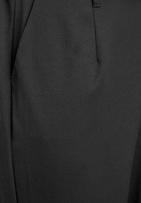 Melnas gludas auduma bikses ar šauru piegriezumu. Tām ir viena priekšējā krokas līnija un slīpi sānu kabatas.