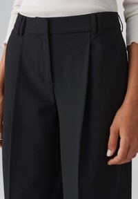 Black pleated trousers with a tailored fit, featuring a smooth texture, angled pockets, and belt loops. Clean lines and minimal detailing.