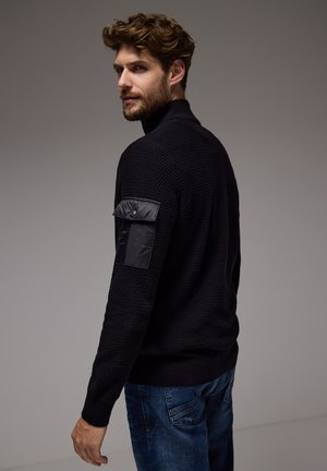 Zwarte geribbelde trui met een hoge kraag, voorzien van een slanke zwarte nylon patch op de linker mouw. Gecombineerd met blauwe denim jeans.