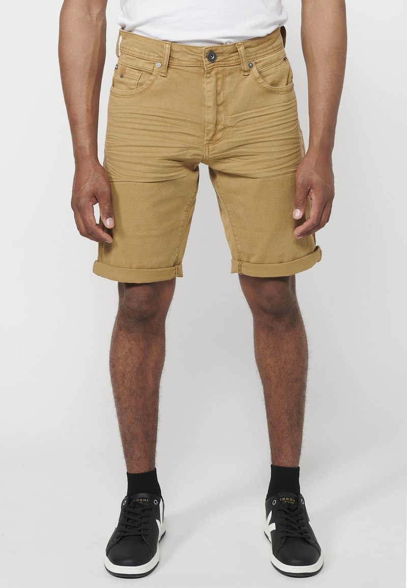 Shorts en denim beige avec un ourlet retroussé, cinq poches et des détails de couture à l'avant. Portés avec des baskets noires et des chaussettes noires.