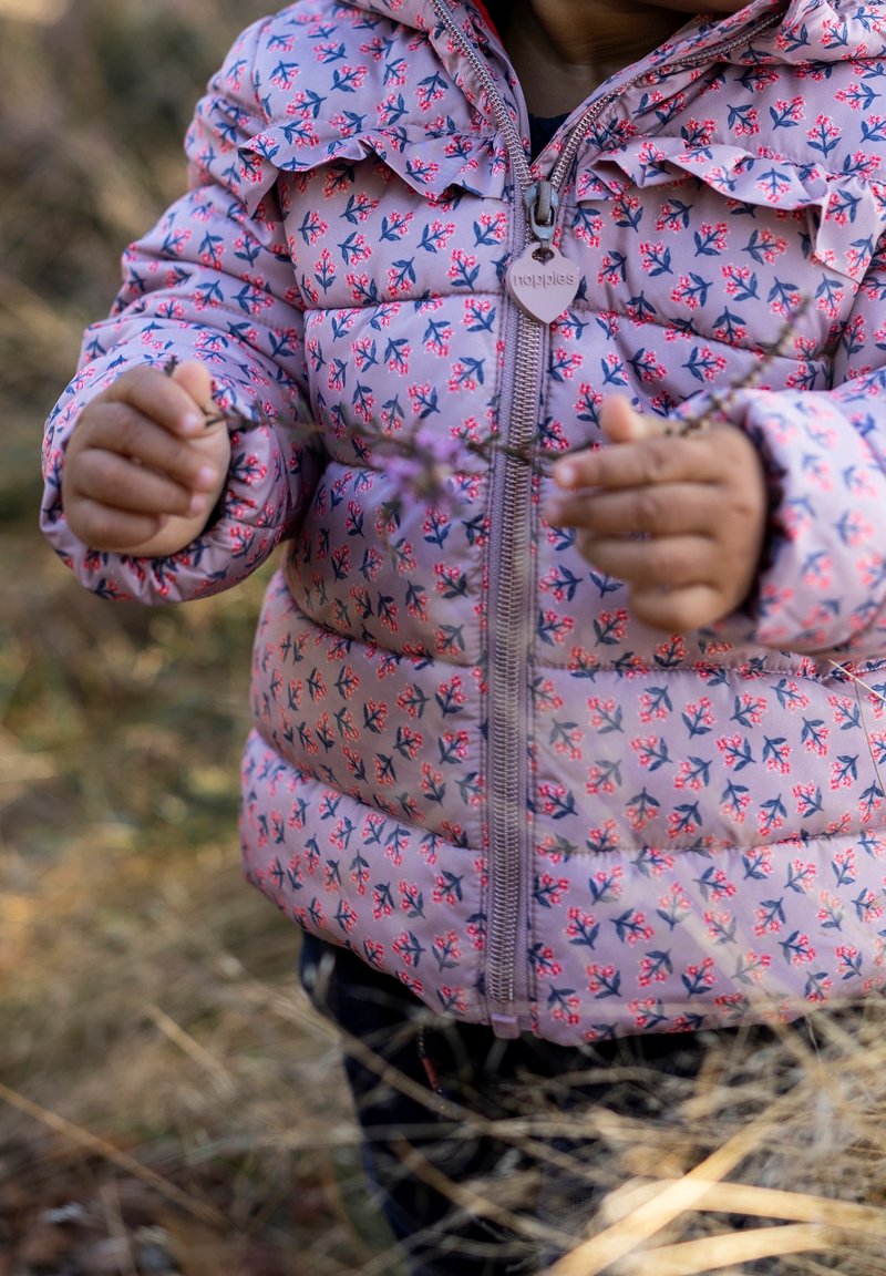 Un enfant portant une doudoune fleurie rose tient de petites fleurs violettes dans les deux mains en plein air, avec de l'herbe sèche en arrière-plan.