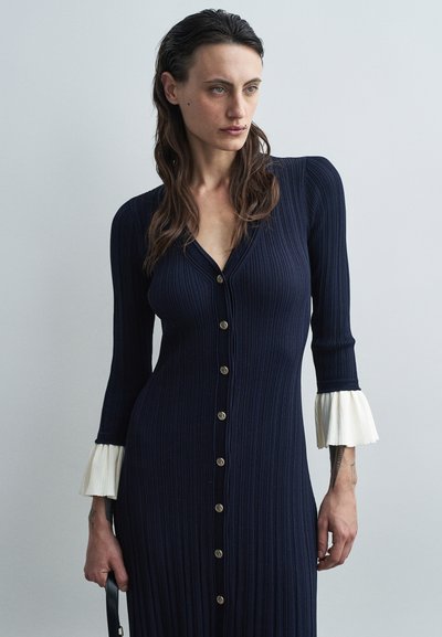 Robe côtelée navy avec un col en V, des boutons dorés et des manches trois-quarts avec des poignets plissés blancs. Texture lisse et silhouette ajustée.