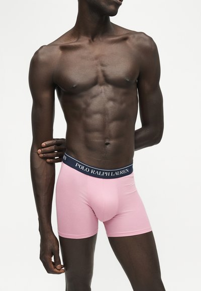 Μυώδης ανδρικός κορμός που φοράει ροζ boxer της Polo Ralph Lauren με ναυτικό μπλε ζώνη, στέκεται μπροστά σε απλό λευκό φόντο.