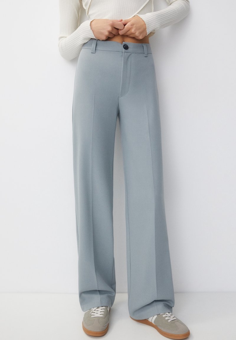 Femme portant un pantalon taille haute bleu clair à jambes larges avec un bouton noir et des baskets grises avec des lacets blancs.