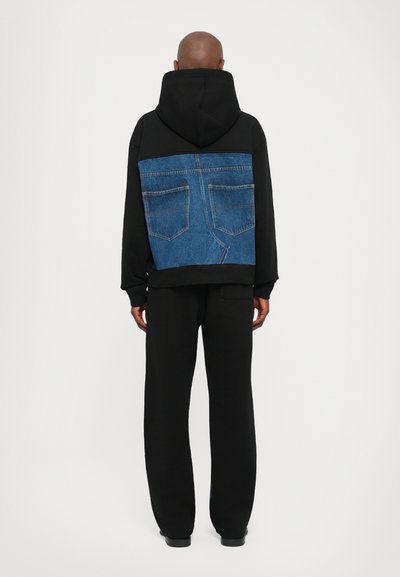 Schwarzer Hoodie mit einem Denim-Patch auf dem Rücken, der ein Design eines Jeansfachs zeigt. Schwarze Jogginghose vervollständigt das Outfit. Minimalistischer Stil.