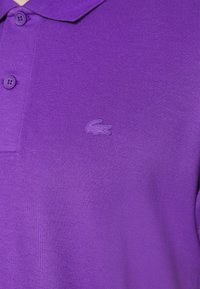 Camisa polo morada hecha de tela texturizada, con una tapeta de dos botones y un pequeño logo en relieve en el lado izquierdo del pecho.