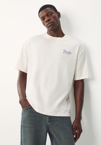 White cotton t-shirt with short sleeves, featuring a purple Fender logo on the left chest. Worn with blue denim jeans, hands in pockets.