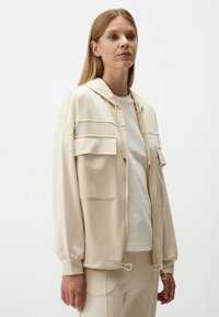 Beige zip-up hoodie met een trekkoordkap, twee voorzakken en geribde manchetten. Zachte stof met een relaxed fit.