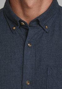 Laivastonsininen napillinen paita, joka on valmistettu teksturoidusta kankaasta. Paidassa on kaulus ja rintatasku, pyöreät ruskeat napit sekä kontrastiväriset ompeleet.