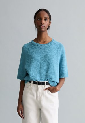 Kobieta ubrana w jasnoniebieski, teksturowany sweter z krótkim rękawem, włożony w białe, wysokowodne spodnie z czarnym paskiem, stojąca na neutralnym tle.