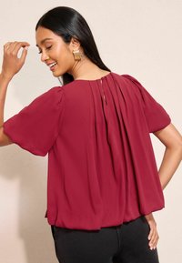 Bordeaux-pusero, jossa on lyhyet puffihihat, laskostettu takaosa ja pieni nappikiinnitys kaula-aukossa. Liukas ja kevyt kangas.