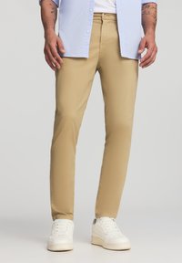 Khaki spodnie slim-fit o gładkiej fakturze i lekkim materiale, w połączeniu z białymi sneakersami i jasnoniebieską koszulą z guzikami.