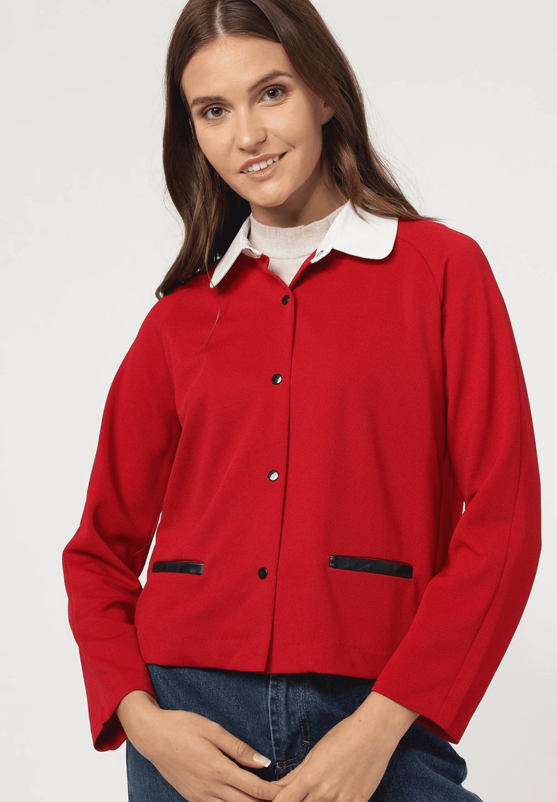 Chaqueta roja con acabado texturizado, botones negros y detalles negros en los bolsillos. Con un collar blanco y mangas largas.