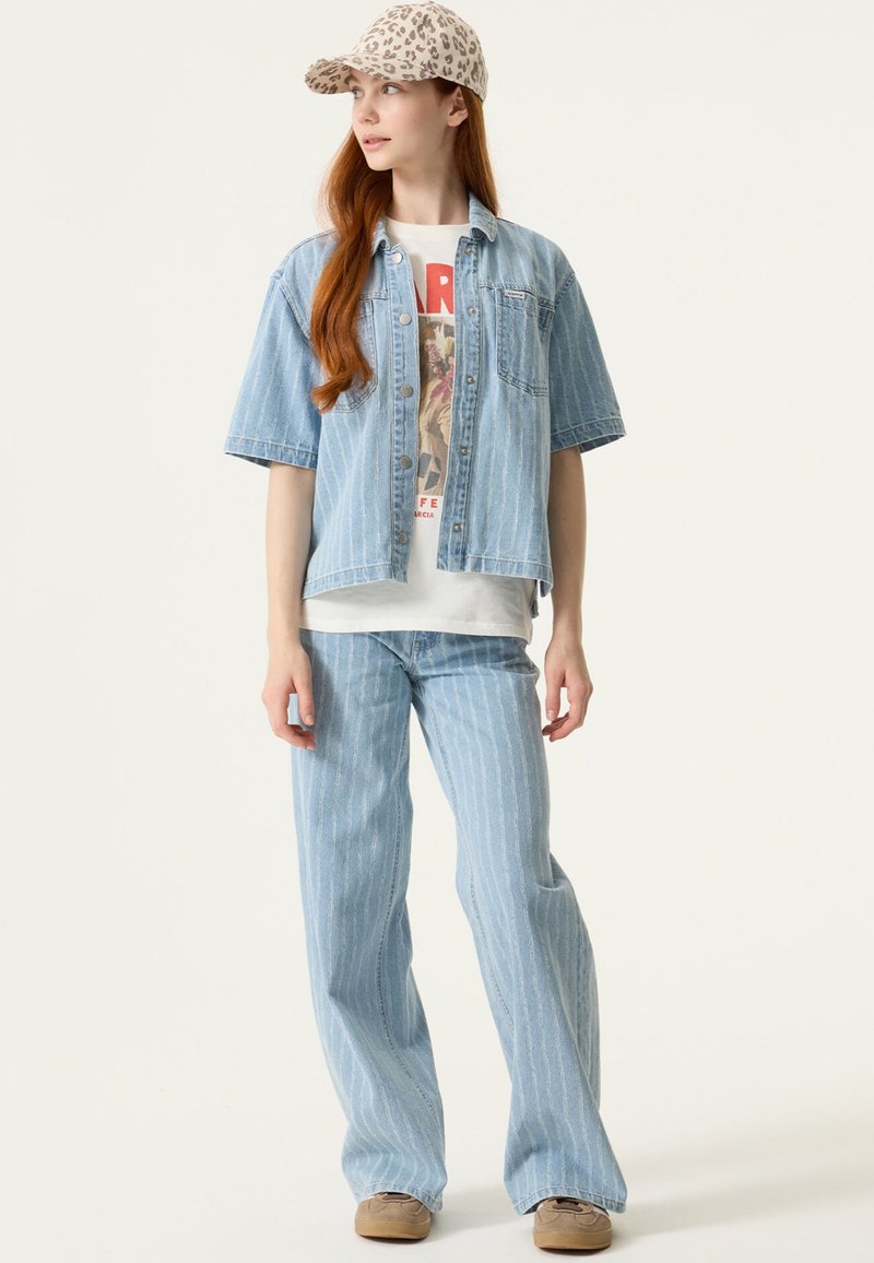 Joven con chaqueta y pantalones de mezclilla azul claro a rayas, camiseta gráfica, gorra de estampado de leopardo y zapatos beige, de pie frente a un fondo blanco.