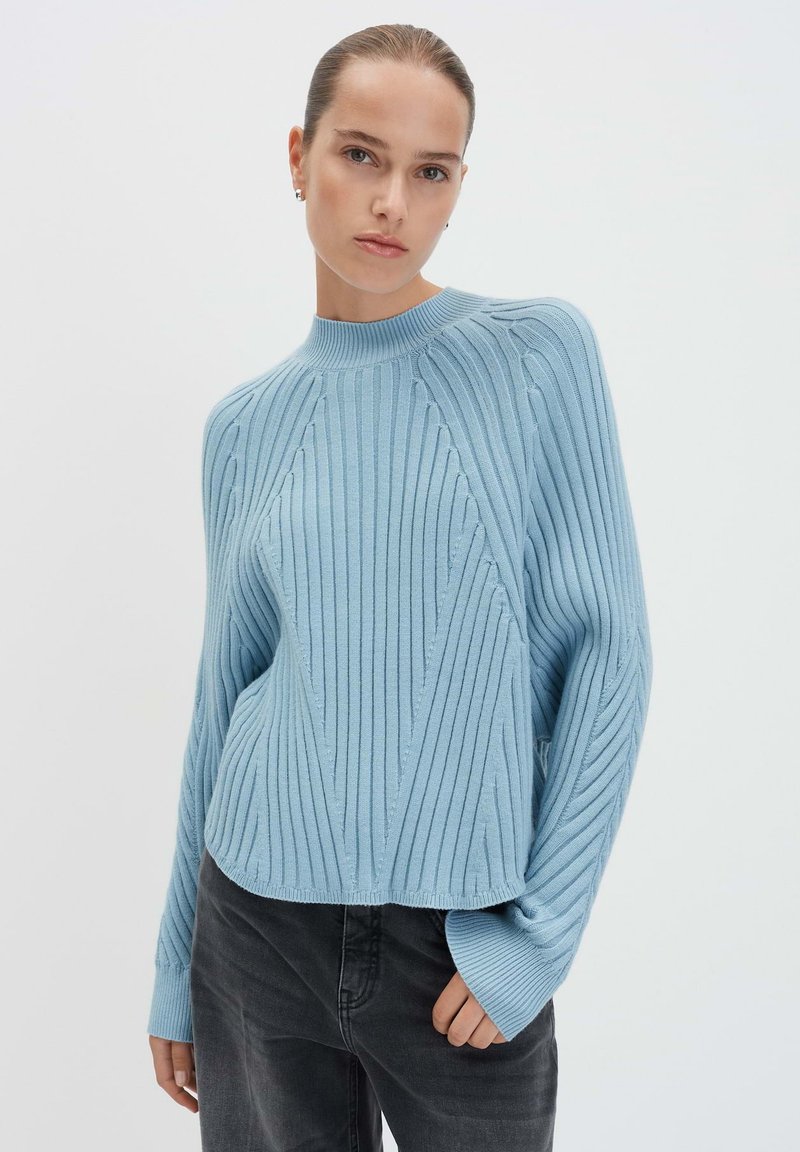 Donna con i capelli all'indietro, che indossa un maglione a coste blu chiaro e pantaloni scuri, in posa davanti a uno sfondo chiaro e semplice.