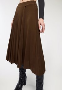 Gonna plissettata marrone realizzata in tessuto morbido, con vita alta e design fluido, abbinata a stivali neri al ginocchio.