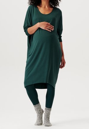 Kobieta w ciąży w ciemnozielonej sukience i legginsach, trzymająca brzuch jedną ręką, stojąca boso w grubych, dzierganych skarpetach.