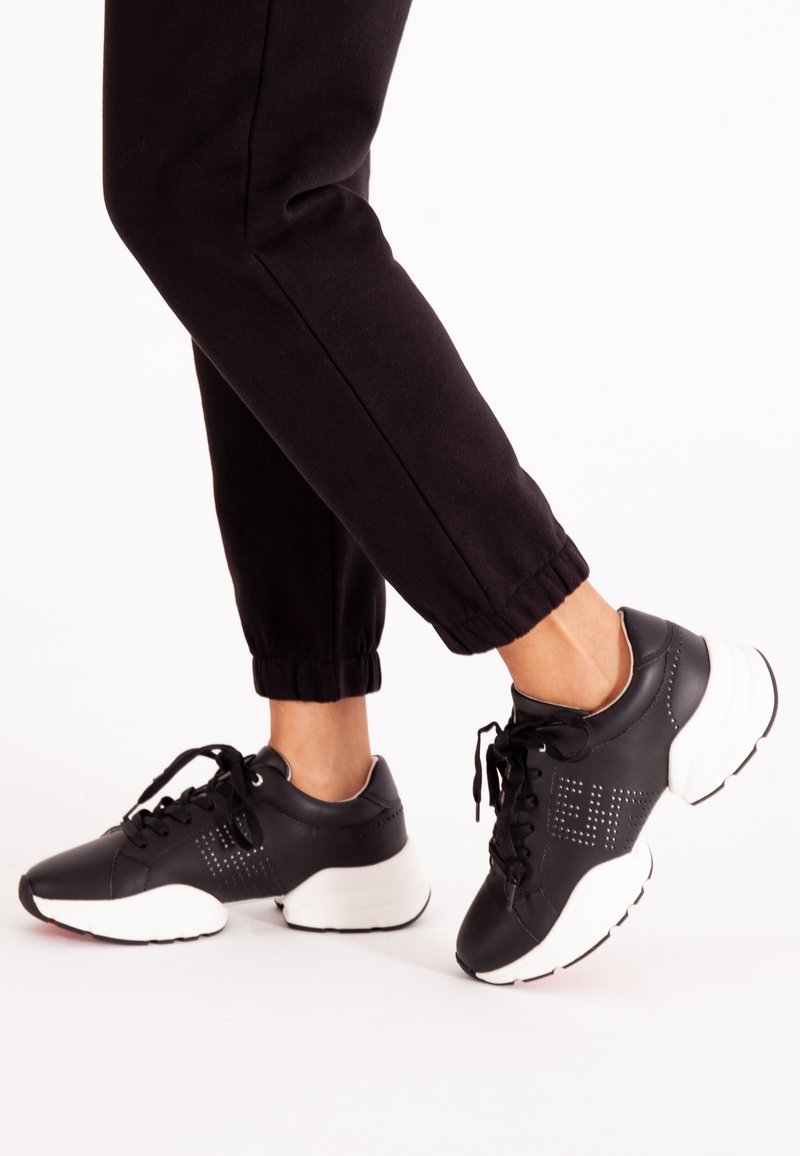 Personne portant un pantalon de jogging noir et des baskets noires avec des semelles blanches épaisses, debout avec un pied légèrement levé sur un fond blanc.