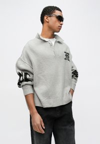 Mladenič s temačnimi sončnimi očali, svetlo sivi široki pleteni pulover z črnimi logotipi in črnimi kavbojkami, stoji z rokami v žepih.