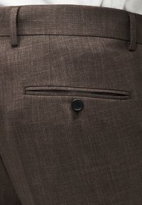 Pantalon habillé en tissu marron texturé avec une poche passepoilée arrière fermée par un bouton noir, porté avec une chemise blanche.