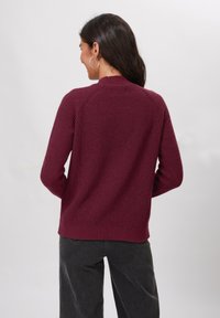 Bordeaux neule, jossa on ribbikuvio, pyöreä kaula-aukko ja raglan-hihat, yhdistettynä tummanharmaisiin housuihin, kuvattu takaa.