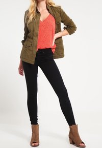 Kaki jacka över en korallfärgad tanktopp med blommönster, kombinerad med svarta skinny jeans och bruna ankelstövlar.