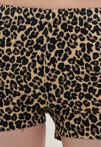 Közelkép egy leopárdmintás rövidnadrágról, amelyet a csípőn és a comb felső részén viselnek, fekete és barna szabálytalan foltokkal bézs háttéren.