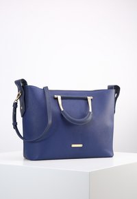 Námořnická modrá umělá kabelka se zlatými detaily, dvojitými držadly a odnímatelným ramenním popruhem. Obsahuje zipové uzávěr a strukturovaný povrch.