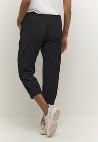 Schwarze, kurze Hosen mit elastischem Bund und Bündchen, aus leichtem Material, kombiniert mit beigen Sportschuhen, die strukturierte Sohlen zeigen.