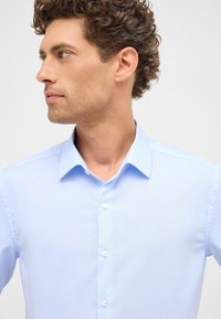Camicia azzurra chiara con colletto, chiusura frontale a bottoni e texture liscia. Presenta chiusure a bottone standard e un design aderente.