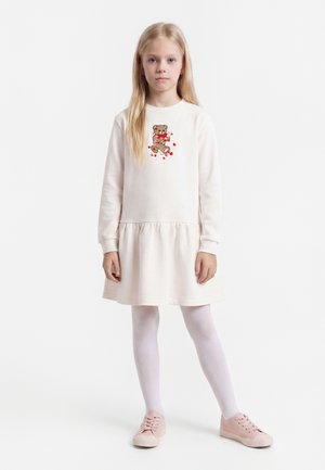 Jeune fille debout portant une robe blanche à manches longues avec un motif d'ours en peluche et des cœurs, des collants blancs et des baskets roses.