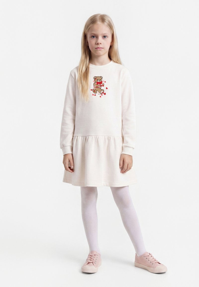 Jeune fille debout portant une robe blanche à manches longues avec un motif d'ours en peluche et des cœurs, des collants blancs et des baskets roses.