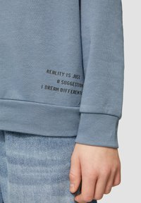 Blauwe katoenen sweatshirt met zwarte tekst op de zoom die zegt "REALITEIT IS VERSCHILLEND, IK DROOM ANDERS." Geribbelde manchetten.