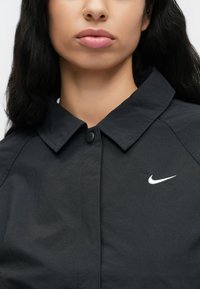 Μαύρο πουκάμισο με κουμπιά από λείο ύφασμα, με μυτερό γιακά και λευκό λογότυπο Nike στην αριστερή πλευρά.