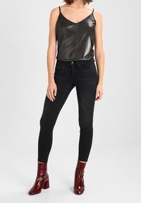 Top sin mangas metálico oscuro con tirantes finos, combinado con jeans negros ajustados y botas de tobillo burdeos brillantes. Textura de tela suave.