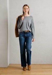 Kvinne iført grå langermet topp, blå jeans og brune støvler, holder en brun jakke over skulderen, står på lys trematrise.