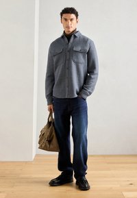 Blue herringbone button-up shirt with two chest pockets, dark jeans, black shoes, and a tan bag, on a model against a neutral background.