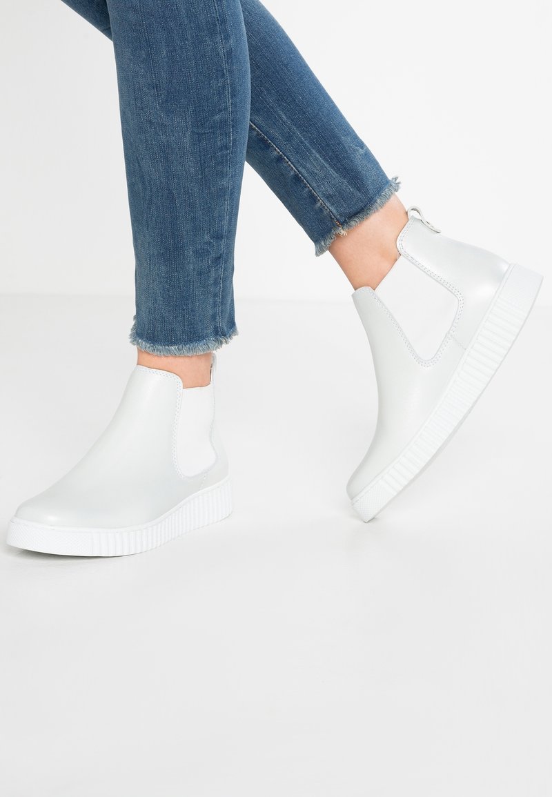 Bottines blanches en matériau lisse avec des panneaux latéraux élastiques, des semelles en caoutchouc texturé et un bout arrondi. Portées avec un jean en denim.