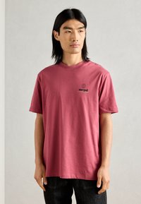 Tricou roz din bumbac, cu guler rotund, mâneci scurte și un mic logo cu glob pământesc și text pe piept. Purtați cu blugi negri.