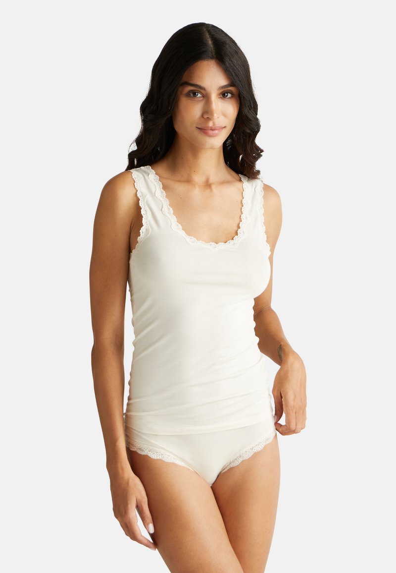 Žena s dlouhými tmavými vlasy, oblečená v krémovém tílku s krajkovým lemem a ladícími spodním prádlem, stojí na světlém pozadí.