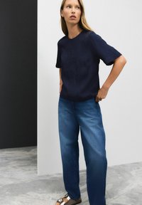 Marineblå kortærmet skjorte med tekstureret mønster, parret med løse blå denim jeans. Brune sandaler med metalliske detaljer fuldender outfittet.