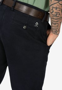 Chinos blu navy in cotone con una tasca sul retro che presenta un design decorativo ricamato bianco. Cintura in pelle marrone con finitura liscia.