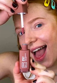 Labio líquido en un envase mate color malva, con un aplicador blanco. La etiqueta muestra "257" y "labio de larga duración". Esmalte de uñas oscuro en los dedos.